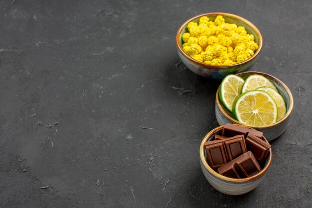 Widok z boku z bliska słodycze w miskach czekoladowe limonki i żółte cukierki w białych miseczkach na ciemnej ścianie