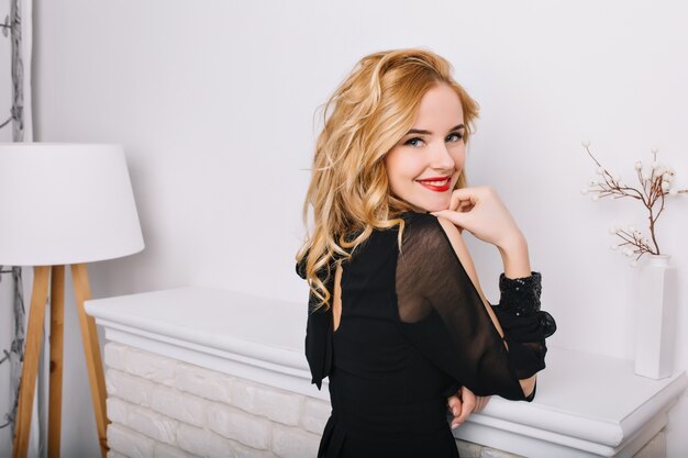Widok z boku zbliżenie portret dziewczyny z blond falowanymi włosami w białym nowoczesnym pokoju z lampą podłogową i kominkiem. Pani uśmiechnięta, pozująca. Ubrana w stylową czarną sukienkę.