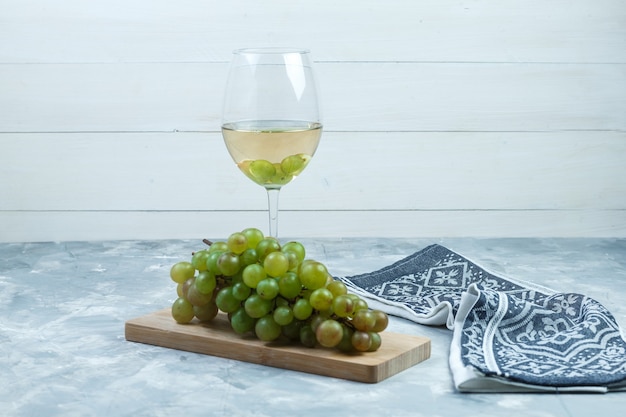 Bezpłatne zdjęcie widok z boku zielone winogrona z lampką wina, kawałek drewna, ręcznik kuchenny na drewnianym i nieczysty szarym tle. poziomy