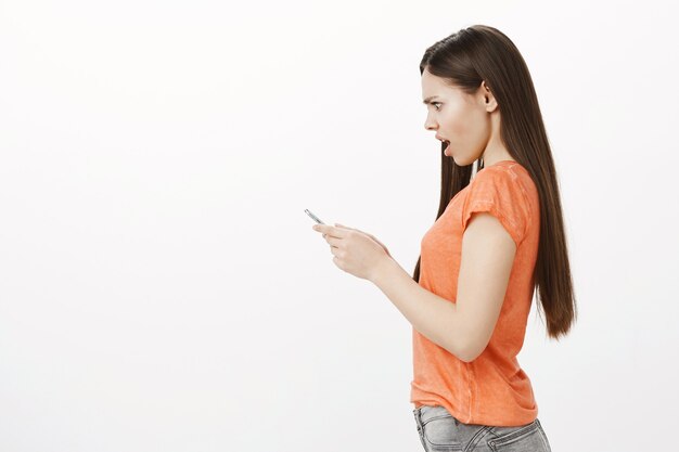 Widok z boku zszokowana i zmartwiona dziewczyna patrzy na ekran telefonu komórkowego z zmartwionym spojrzeniem