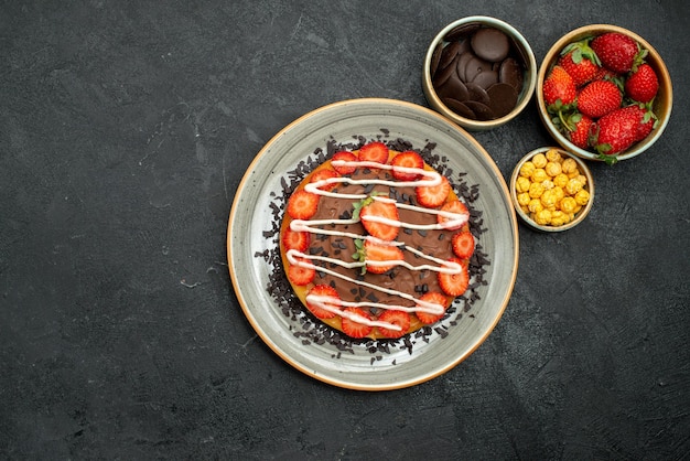Widok z góry apetyczny tort z kawałkami czekolady i truskawek na białym talerzu i miski z czekoladową truskawką i orzechami laskowymi na środku ciemnego stołu