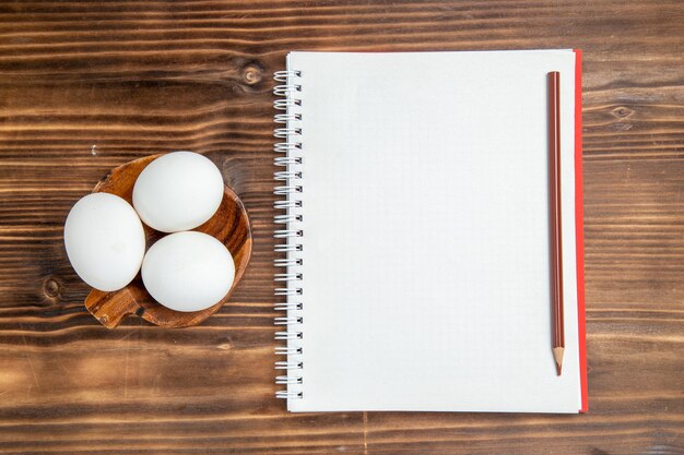 Widok z góry całe surowe jajka z notatnikiem na brązowej powierzchni drewnianej posiłek jedzenie śniadanie drewno