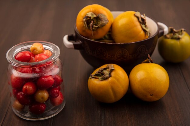 Widok z góry czerwonych wiśni derenia na szklanym słoju z owocami persimmon na misce na drewnianej powierzchni