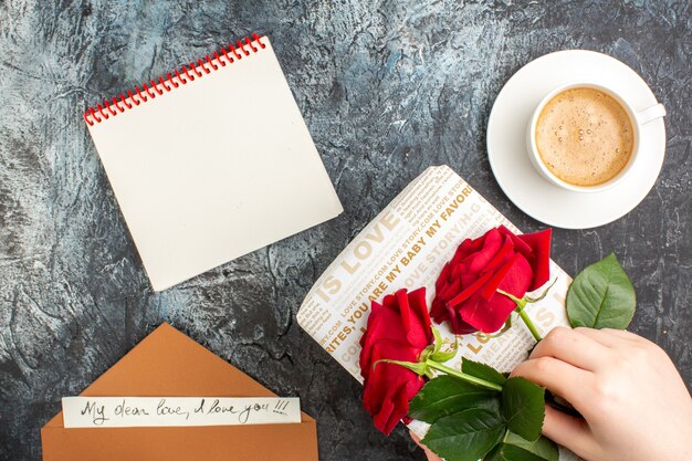 Widok z góry dłoni trzymającej czerwone róże na pięknym pudełku i filiżance kawy w kopercie z notatnikiem z listem miłosnym na lodowatej ciemnej powierzchni