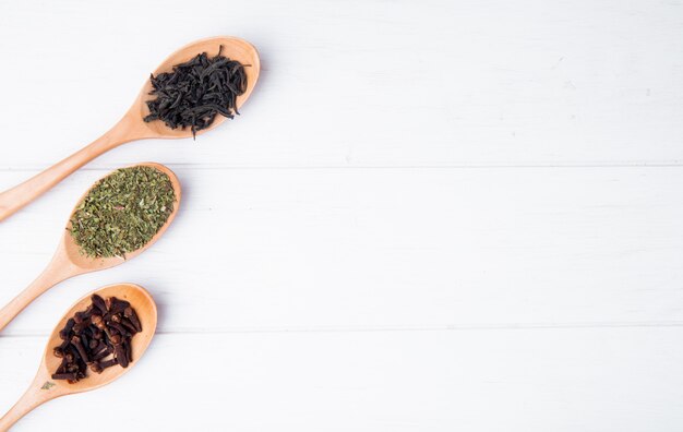 Widok z góry drewniane łyżki z przyprawami i ziołami suszą liście czarnej herbaty, przyprawy goździkowe i suszoną miętę na białym drewnie z miejsca kopiowania