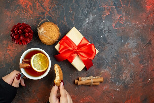Bezpłatne zdjęcie widok z góry filiżanka herbaty o smaku cytryny i cynamonu i ciastko w kobiecej dłoni prezent świąteczny czerwony pinecone laski cynamonu ciasteczka na ciemnoczerwonym stole