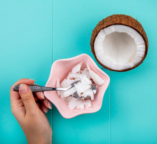 Widok z góry kobiecej ręki trzymającej łyżkę z pulpy kokosowej w różowej misce z przepołowionym kokosem na niebiesko