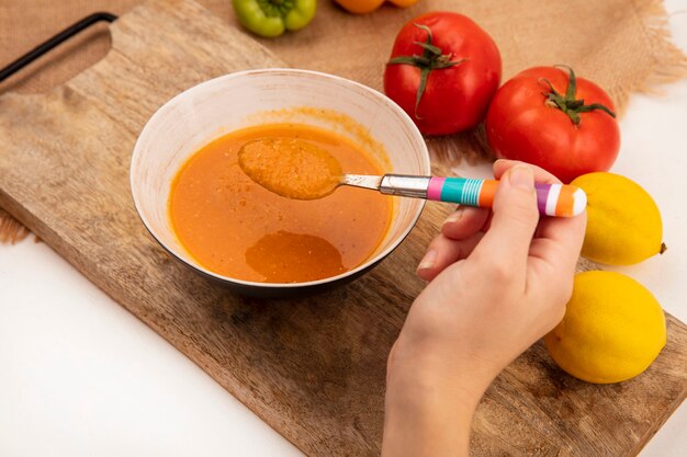 Widok z góry kobiecej ręki trzymającej łyżkę z zupą z soczewicy na misce na drewnianej desce kuchennej na woreczku z cytrynami i pomidorami odizolowanymi na białej powierzchni