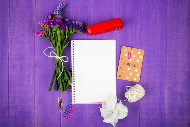 Widok z góry kwiaty statice w kolorze fioletowym i różowym z szkicownikiem czerwony zszywacz liny i pocztówka na fioletowym tle drewnianych