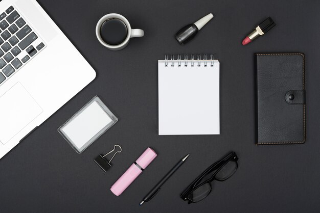 Widok z góry laptopa z filiżanką kawy; szminka; lakier do paznokci i rzeczy biurowe na czarnym tle