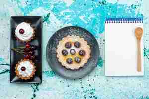 Bezpłatne zdjęcie widok z góry małe ciasteczka ze śmietaną i owocami wraz z notatnikiem na jasnoniebieskim stole do pieczenia owoców