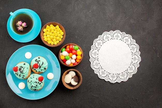 Widok z góry małe kremowe ciasta z filiżanką herbaty i cukierków na ciemnej powierzchni kolor deseru z kremem herbacianym