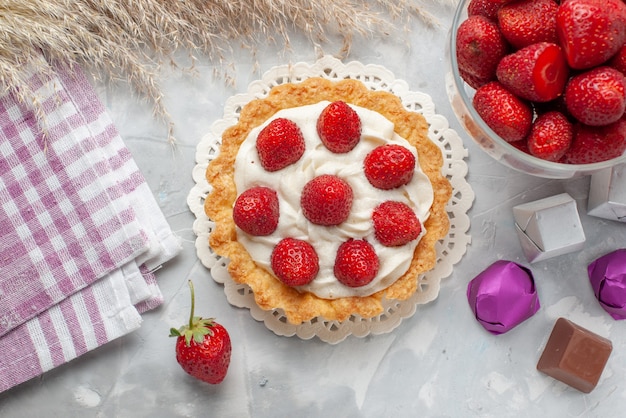 Widok z góry małe kremowe ciasto ze świeżymi czerwonymi truskawkami i czekoladowymi cukierkami na białym oświetleniu na biurku ciasto owocowo-jagodowe biszkoptowe