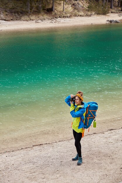 Widok z góry na aktywną kobietę stojącą nad brzegiem turkusowego górskiego jeziora, strzela coś do aparatu, nosi dużą torbę