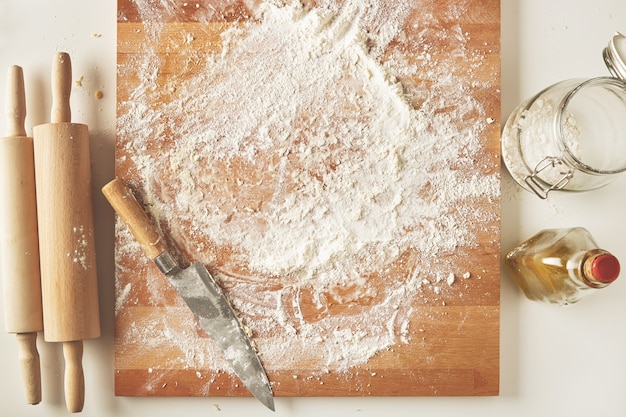 Widok z góry na biały stół z izolowaną drewnianą deską z nożem, dwoma wałkami do ciasta, butelką oliwy z oliwek, przezroczystym słojem z mąką. Prezentacja procesu gotowania