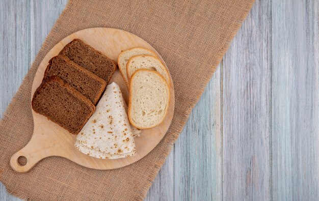 Widok z góry na chleb jako pokrojone białe żytnie i podpłomyki na desce do krojenia na worze na drewnianym tle z miejscem na kopię