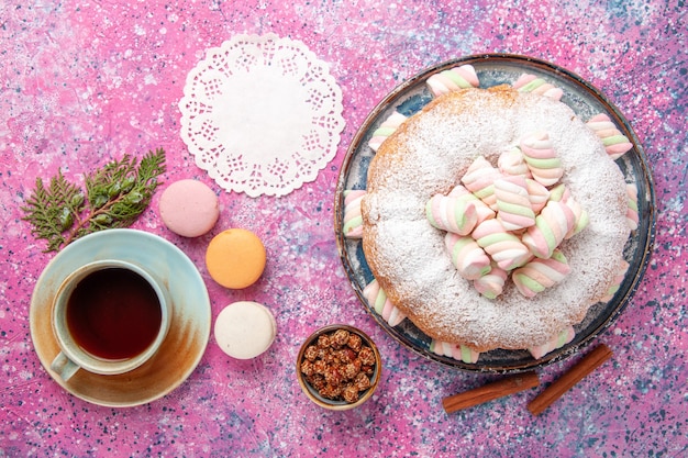 Widok z góry na ciasto cukrowe w proszku z filiżanką herbaty i francuskimi makaronikami na różowym biurku