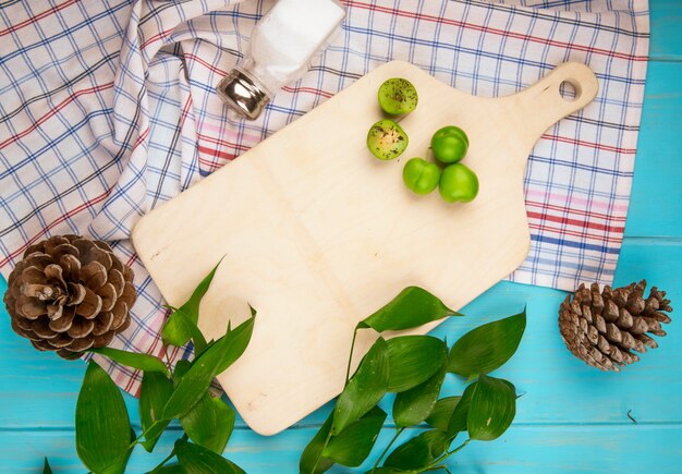 Widok z góry na drewnianą deskę i rozrzucone kwaśne zielone śliwki ze stożkami na kraciastej tkaninie na niebieskim drewnianym stole