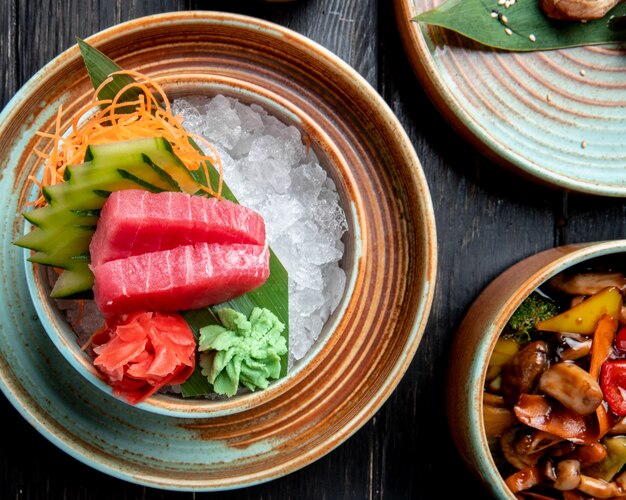 Widok z góry na kawałek sashimi z tuńczyka z ogórkami, imbirem i sosem wasabi na kostkach lodu w misce na drewnianym stole