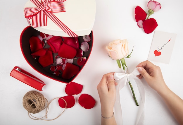 Widok z góry na pudełko w kształcie serca wypełnione czerwonymi płatkami róży lina zszywacza w kolorze czerwonym i kobiece ręce wiążące białą różę wstążką na białym tle