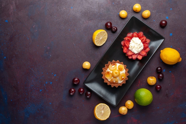 Widok z góry na pyszne kremowe ciasta wewnątrz płyty ze świeżymi cytrynami i owocami na ciemnej podłodze ciasto owocowe herbatniki słodkie wypieki