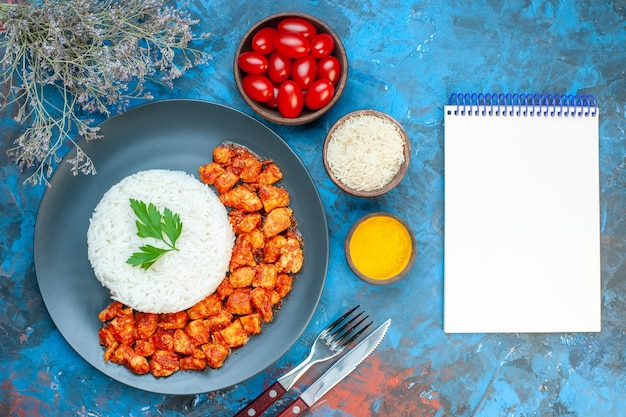 Widok z góry na pyszny posiłek ryżowy z zielonym i pomidorowym zestawem sztućców do kurczaka sól pomidory z czosnkiem obok notebooka na niebieskim tle