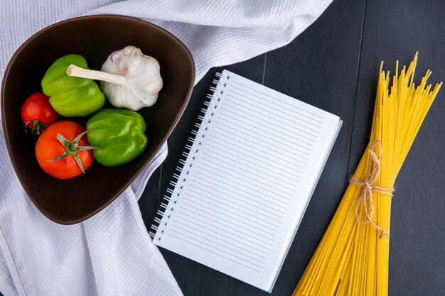 Widok z góry na surowe spaghetti z pomidorami, czosnkiem i papryką w misce na białym ręczniku i notatniku na czarnej powierzchni