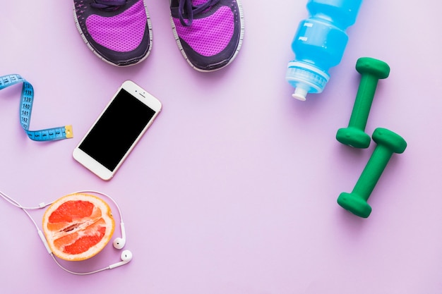Widok z góry na taśmę pomiarową; hantle; buty; o połowę pomarańczowe owoce; butelka wody; telefon i słuchawki na różowym tle