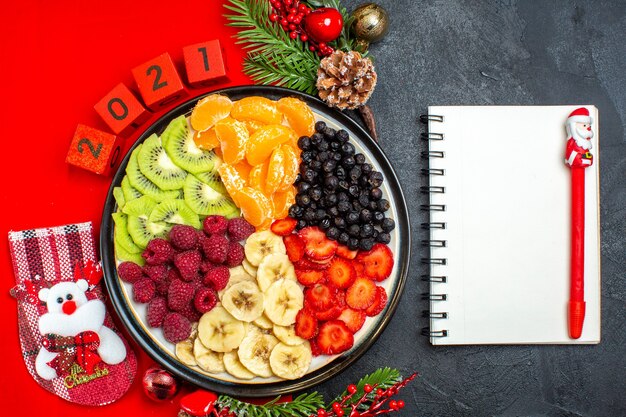 Widok z góry na zbiór świeżych owoców na talerzu obiadowym akcesoria do dekoracji gałązki jodły i numery świąteczne skarpety na czerwonej serwetce następny notatnik z piórem na czarnym tle