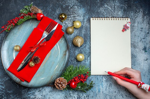 Widok z góry na zestaw sztućców z czerwoną wstążką na ozdobnej serwetce na niebieskim talerzu i akcesoria świąteczne ręka trzyma długopis na notebooku na ciemnym tle