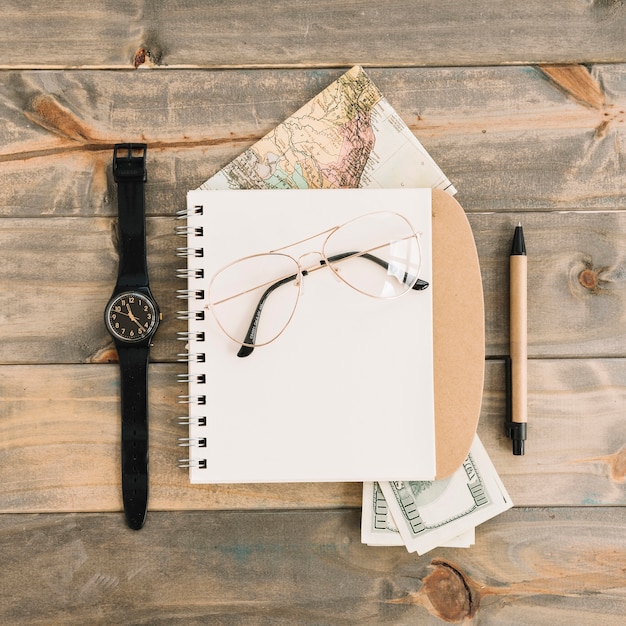Widok z góry okularów na spirali notatnika; waluta; mapa; zegarek i długopis na tle drewnianych desek