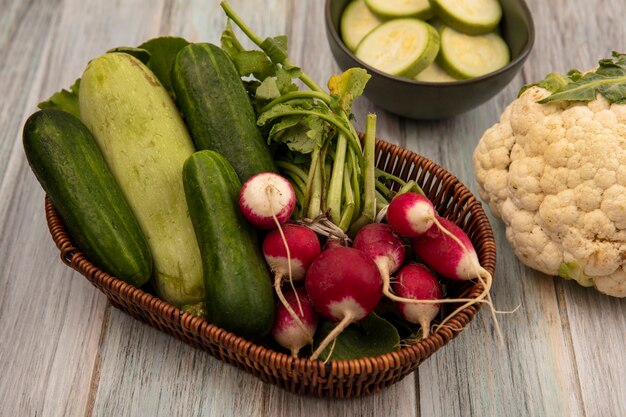 Widok z góry organicznych warzyw, takich jak ogórki, cukinia i rzodkiewki, na wiadrze z kalafiorem na szarym tle drewnianych