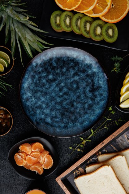 Widok z góry owoców cytrusowych jako ananasa kiwi pomarańcza limonka plasterki cytryny i mandarynki z kromkami chleba na tacy i dżemem w misce wokół pustego talerza na czarnym tle