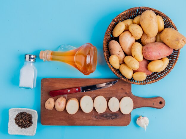 Widok z góry plastry ziemniaków i nóż na desce do krojenia z całych w koszyku masła sól i czarny pieprz i czosnek na niebieskiej powierzchni