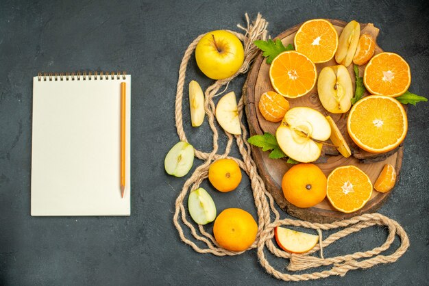 Widok z góry pokrojone jabłka i pomarańcze na desce koktajlowej zeszyt i ołówek na ciemnym tle