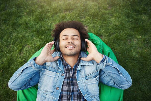 Widok z góry portret zadowolony, zrelaksowany Afroamerykanin z włosiem leżącym na trawie, słuchając muzyki z zamkniętymi oczami i uśmiechem, będąc szczęśliwym i ciesząc się dźwiękami w parku