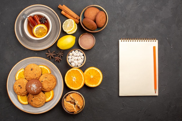 Widok z góry pyszne piaskowe ciasteczka ze świeżymi pomarańczami i filiżanką herbaty na ciemnym tle owoce herbatniki słodkie ciastko cukier cytrusowy