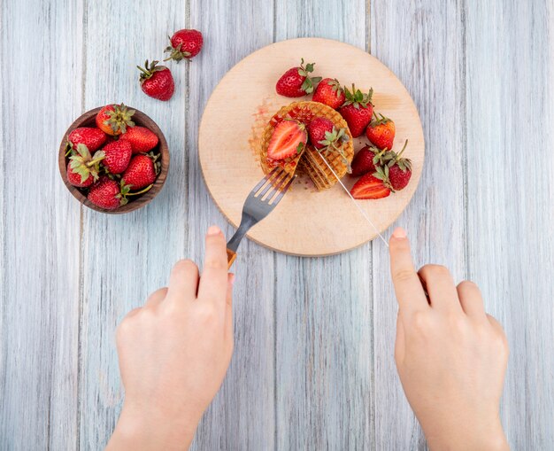 Widok z góry rąk cięcia ciastek wafel widelcem i nożem na deskę do krojenia i miska truskawek na powierzchni drewnianych