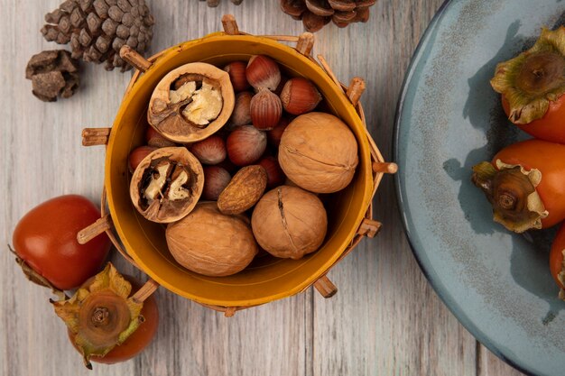 Widok z góry różnych orzechów, takich jak orzechy włoskie i orzechy laskowe, na wiadrze z miękkimi świeżymi persimmons na talerzu na szarej drewnianej ścianie