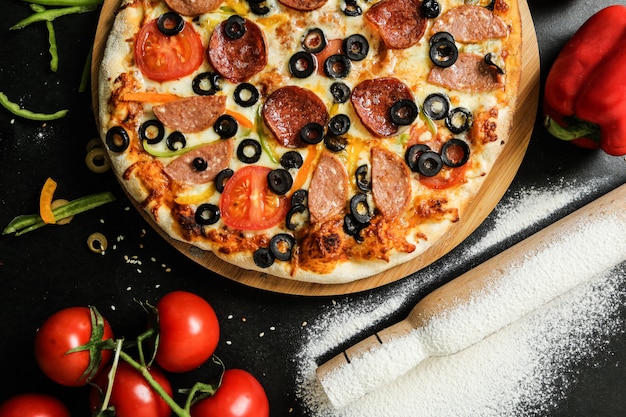 Widok z góry salami pizza z pomidorami, papryką i oliwkami na tacy z mąką i wałkiem do ciasta