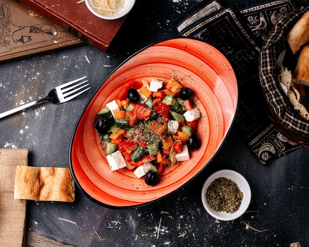 Widok z góry Sałatka grecka świeża bogata w witaminę Yummy z pokrojonymi warzywami wewnątrz czarnego talerza i chlebem na ciemnej powierzchni