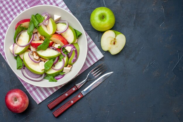 Widok z góry sałatka ze świeżych jabłek na okrągłym talerzu zielone i czerwone jabłka nóż i widelec na ciemnym stole