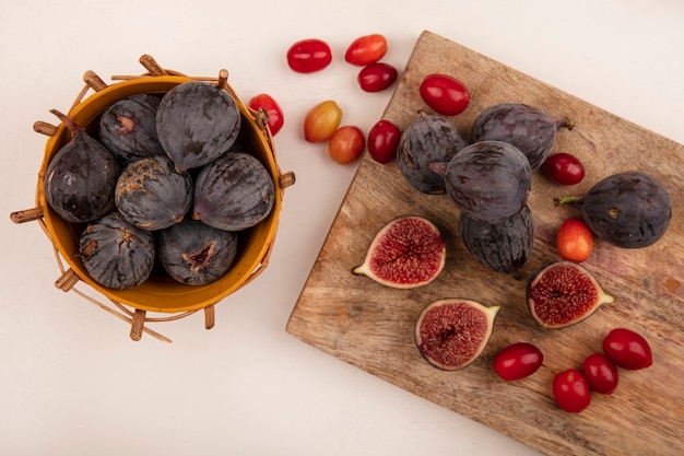 Bezpłatne zdjęcie widok z góry słodkich czarnych fig na wiadrze z czarnymi figami i wiśniami dereń na drewnianej desce kuchennej na białej ścianie