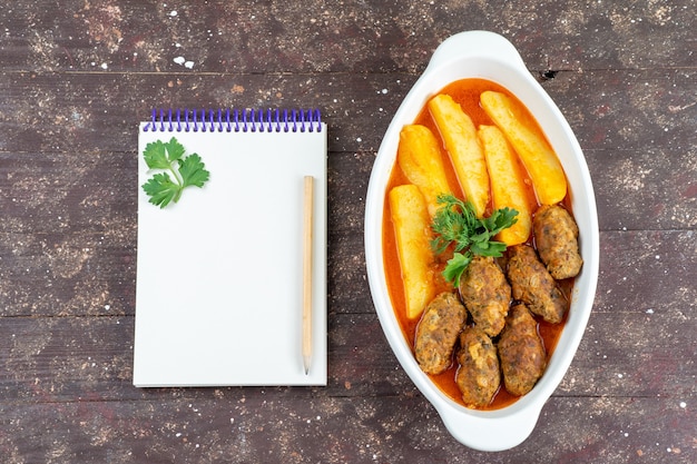 Widok z góry smaczne kotlety mięsne gotowane wraz z ziemniakami i sosem wewnątrz talerza z notatnikiem na brązowym biurku danie ziemniaczane danie mięsne obiad