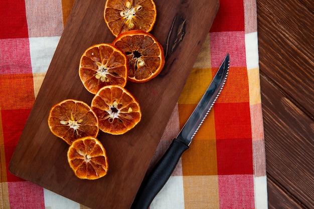 Bezpłatne zdjęcie widok z góry suszonych plasterków pomarańczy z kuchennym nożem na drewnianej desce do krojenia na kraciastym obrusie