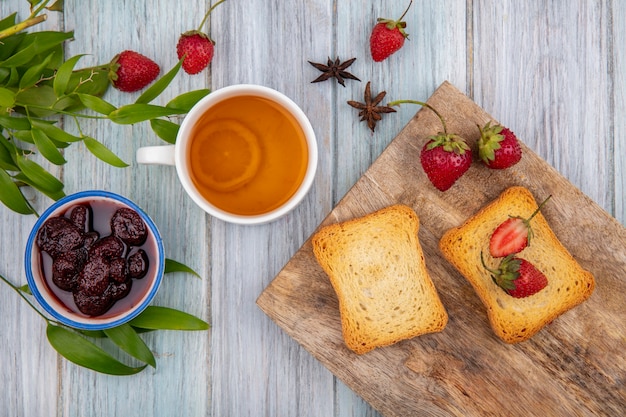 Widok Z Góry świeżych Truskawek Na Drewnianej Desce Kuchennej Z Tostami Kromkami Chleba Z Dżemem Truskawkowym Z Filiżanką Herbaty Na Szarym Drewnianym Tle