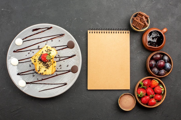 Bezpłatne zdjęcie widok z góry tort z notatnikiem z kremem truskawkowym między truskawkami czekolada w miseczkach i talerzem ciasta z sosem czekoladowym na ciemnej powierzchni