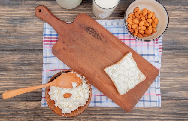 widok z góry twarogu z drewnianą łyżką w misce i migdałów mlecznych w misce z kromką chleba na desce do krojenia na kratce i drewnianym stole