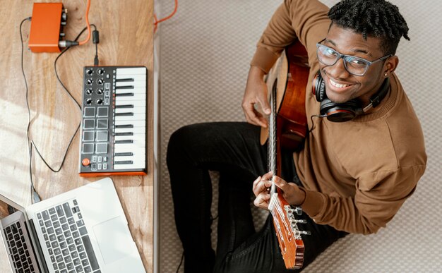 Widok z góry uśmiechniętego muzyka płci męskiej w domu, gra na gitarze i miksowanie z laptopem