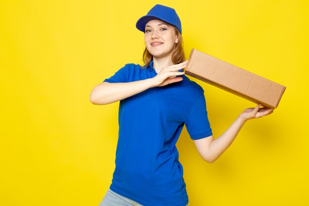 Widok z przodu atrakcyjna kobieta kurier w niebieskiej koszulce polo niebieskiej czapce i dżinsach, uśmiechając się, śpiesząc się, trzymając pakiet na żółtym tle usługi gastronomicznej